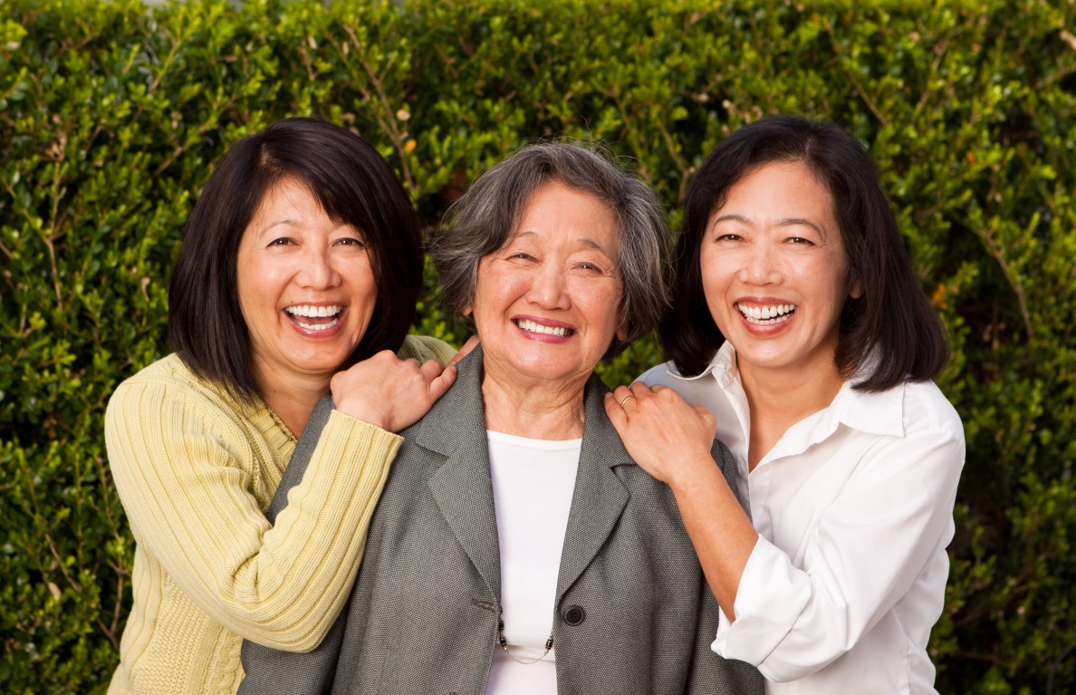 An elderly mother with her two adult daughters, smiling outdoors in front of a green hedge.